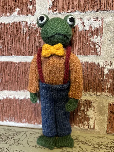 Hand-knitted green frog wearing a mustard cardigan and yellow bow tie