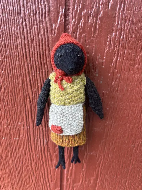 Hand-knitted black bird wearing a cream apron and red headscarf